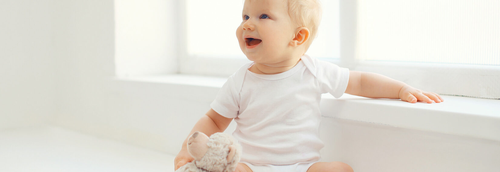 Baby sitzt in einem weißen Raum in der nähe des Fensters. Es hat einen Teddybären bei sich.