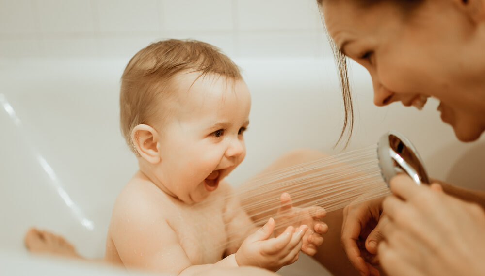 Ein Baby sitzt in der Badewanne und wird mit dem Duschkopf von seiner Mama abgeduscht.