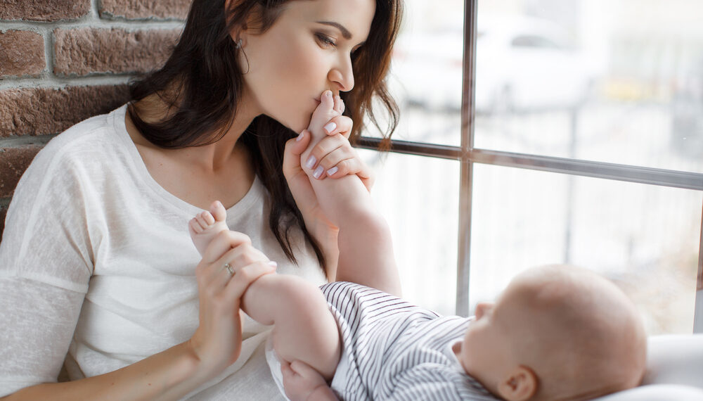 Die Mutter hat das Baby auf dem Schoß. Das Baby liegt mit dem Rücken auf den Beinen der Mutter. Die Mutter küsst den einen Fuß des Babys.