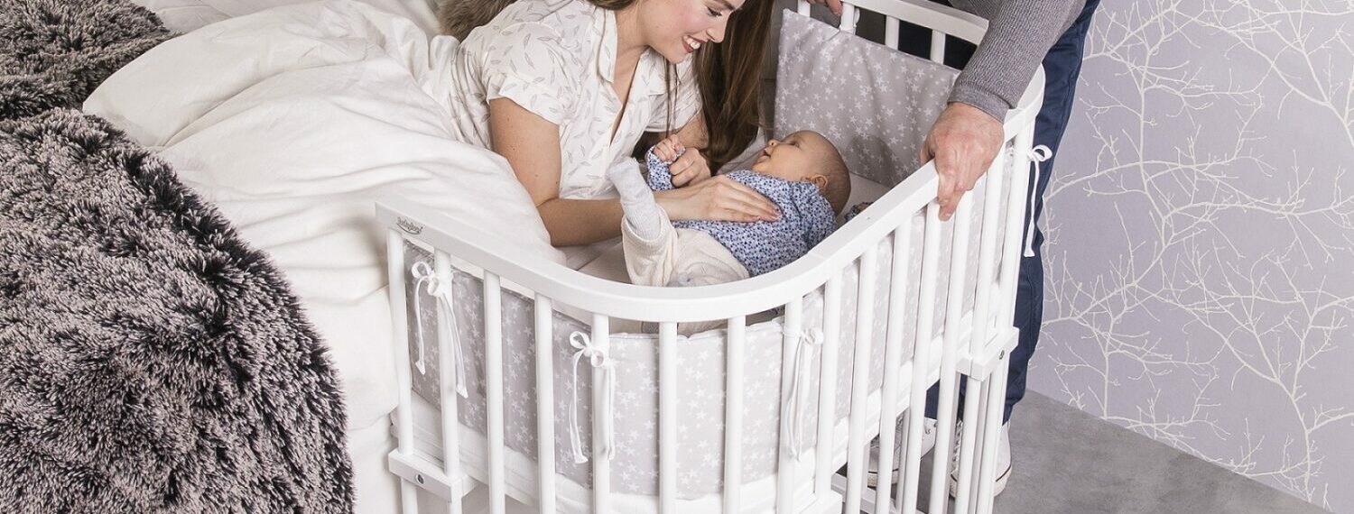 Mutter liegt im Bett, das Baby liegt im Beistellbettchen daneben. Der Vater und die Mutter beugen sich beide zu dem Kind.