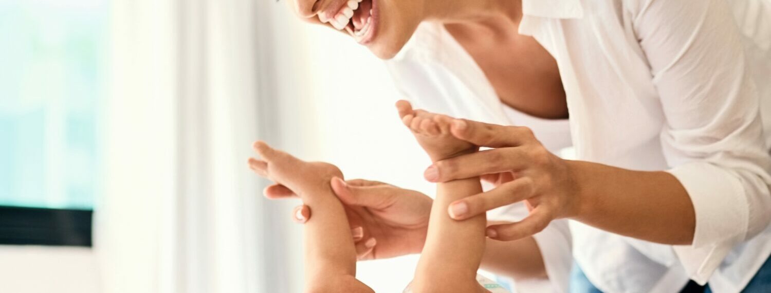 Lachende Mutter beugt sich über ihr auf dem Rücken liegendes Baby und fasst an die Füße