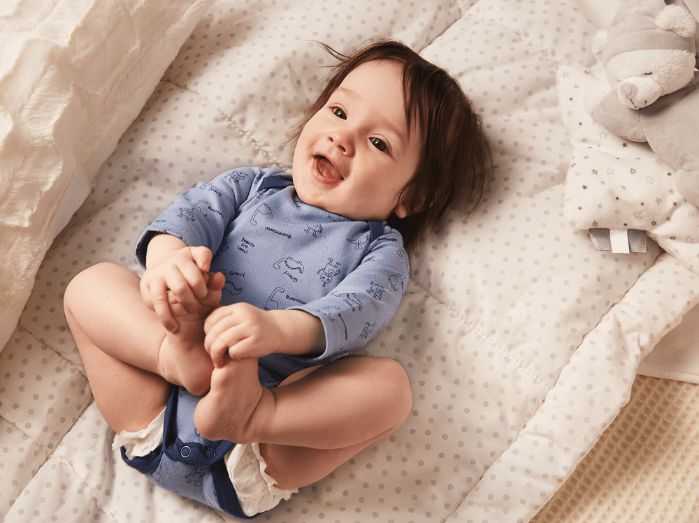 Lachendes Baby mit C&A Baby Body liegt lachend auf dem Rücken und fasst sich an die Füße