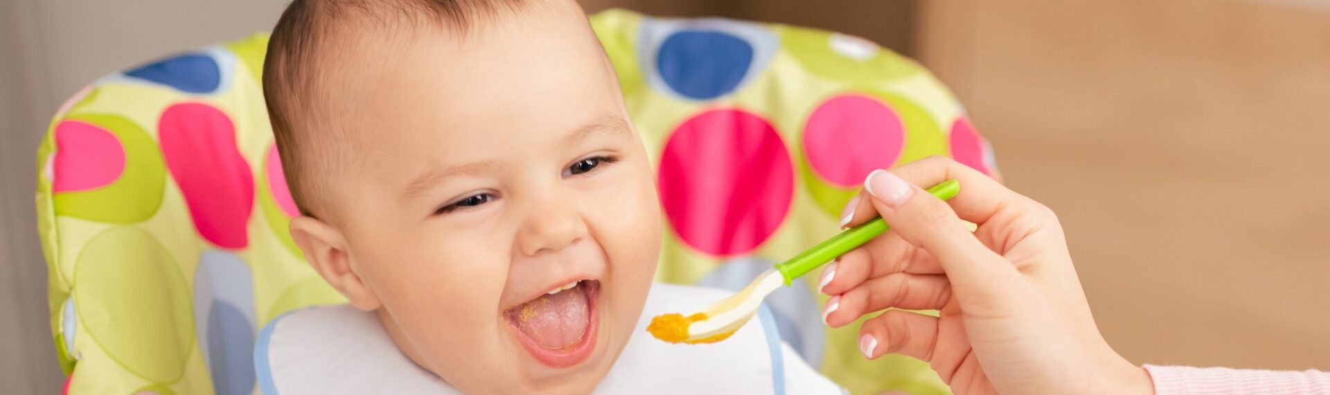 1. Eine Mutter füttert ihr Baby mit einem Löffel, während das Baby zufrieden aussieht. Dieses Bild veranschaulicht die Einführung von fester Nahrung im ersten Lebensjahr.